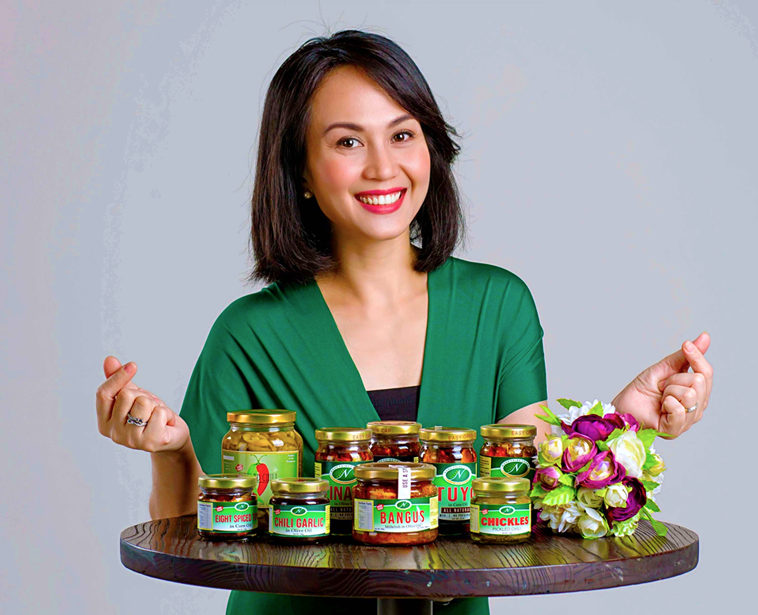 Nene Tamayo, owner of Nene Prime Foods, smiling and flashing finger hearts over a table full of their best-selling products